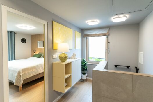 Dielenbereich im Obergeschoss: heller Fliesenboden, schlanke Wandkonsole mit integriertem Stauraum und gelben Kunstakzenten. Tageslicht strömt durch das Fenster, von hier führen Türen in vier separate Schlafzimmer und die Treppe ins Erdgeschoss.