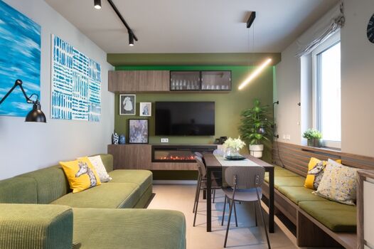 Gemütliches Wohn-Esszimmer mit grünem Cord-Sofa und passender Bank, eleganter Esstafel für 8 Gäste, modernem Elektro-Kamin unter dem 55'' Smart-TV, stimmungsvoller LED-Leuchte und liebevoll ausgewählten Kunst- und Pflanzenakzenten.