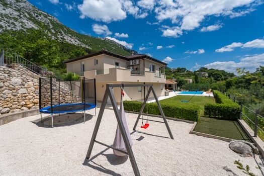 Umzäunter Spielplatz mit Trampolin, Schaukel, Badmintonplatz, Rasenminigolf und Lounge-Sitzbereich